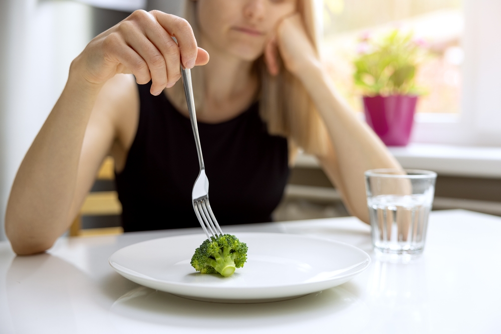 woman looking sad using a fork to play with a piece of broccoli on her plate