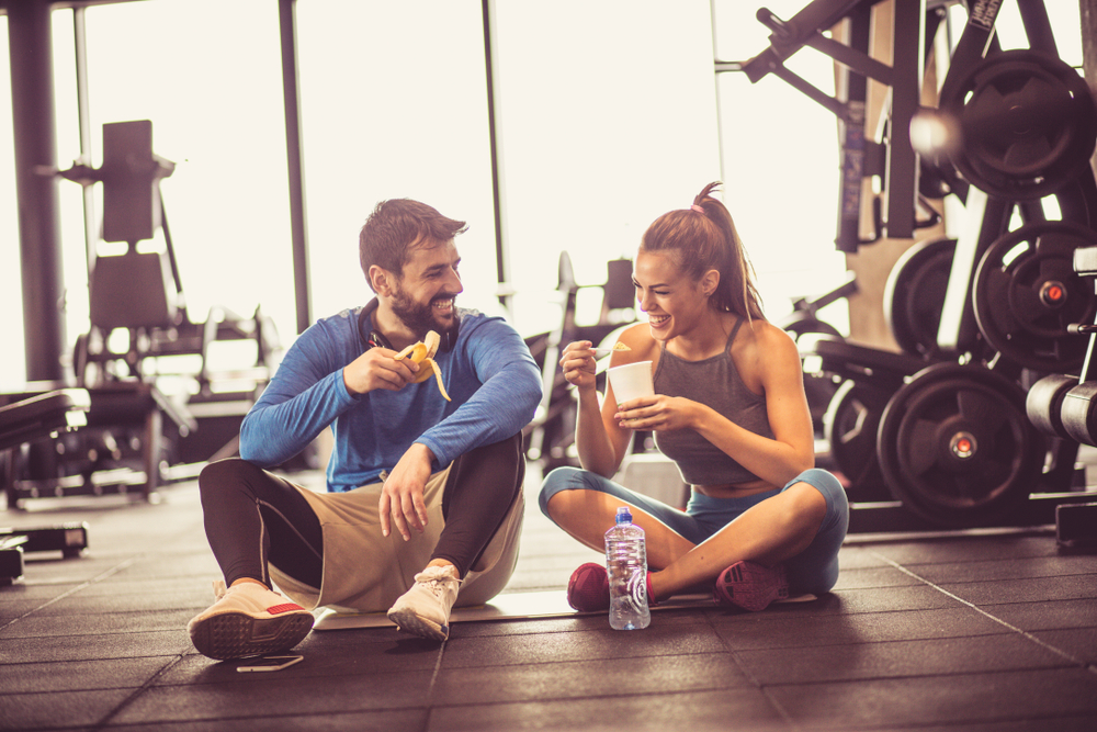 man and woman at gym smiling, eat snacks on the floor